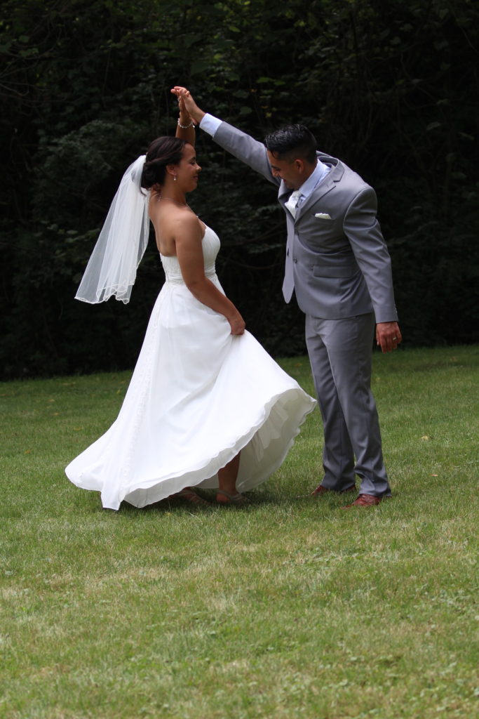 Bride in a wedding dress being spun by the groom in a grey suit. They are outside on grass with a wall of trees in the background