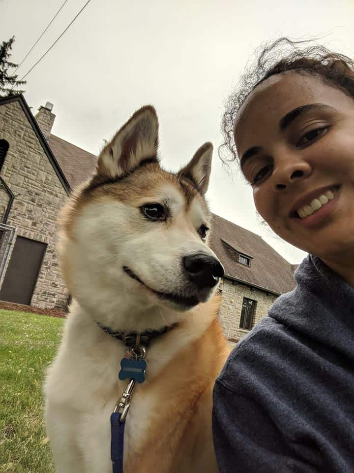 A close-up of a woman's face and her dog (a red and white Shiba Inu mix) in a park. There is a brick building in the background.