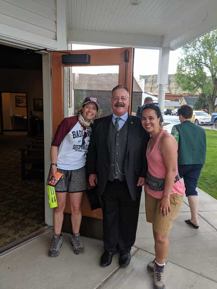 A Theodore Roosevelt impersonator dressed in a dark suit is standing in the middle of two women. All are smiling at camera. Woman on the right is wearing  a 3/4 length sleeve shirt (white body, maroon sleeves), grey shorts and a maroon baseball cap. Women on right is wearing pink sports tank tops and khaki shorts.