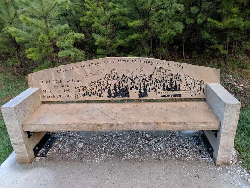 Bench on the Black Elk Peak trail. The bench is made of stone. The back of the bench has a sketch of the mountain with trees and a quote at the top "Life is a journey, take time to enjoy every step"