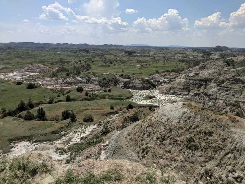 View of Theodore Roosevelt National Park. The right side of the frame are mountains with trees spotted throughout them but they are mostly grey in color. The left side of the frame is more flat with grass and more trees dotted throughout the landscape.