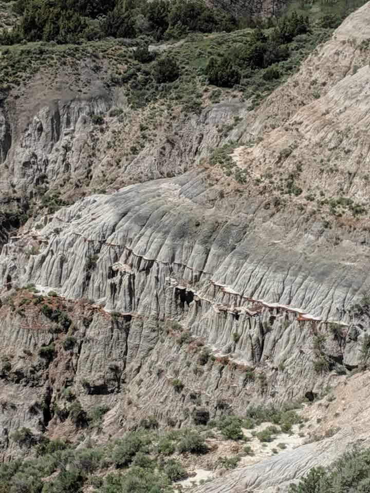 Close up of the side of a mountain. The mountain is made up of rings of colors in hues of grey, browns, whites, and reds. Some rings looked like they draped over the ring beneath them like a skirt. There are bushes and trees dotted along the mountain.