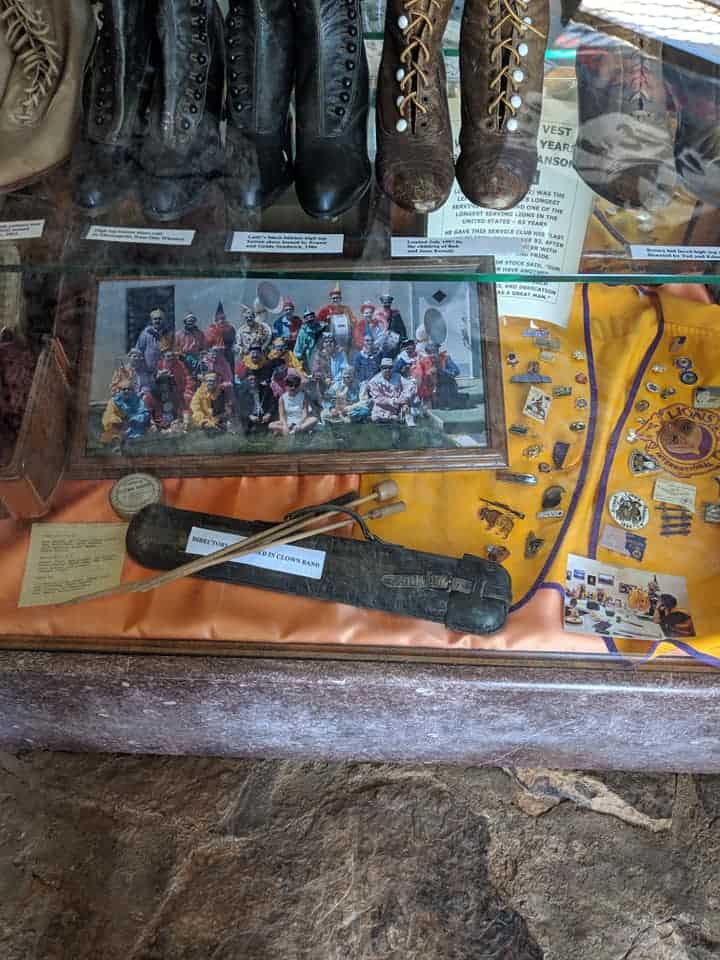 2 shelves in a display case in the museum at Petrified Wood Park. The top shelf has several pair of old boots. The bottom shelf has a picture of clowns that are member of a band and a conductor's wand.