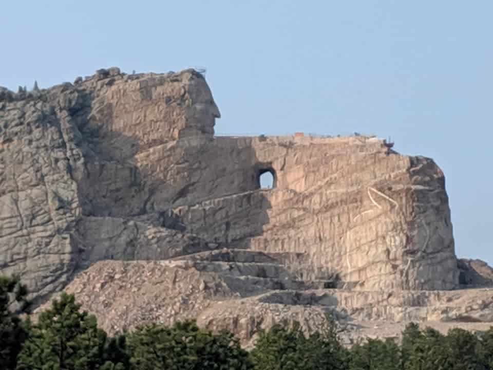Crazy Horse Memorial. The face has been chiseled out from the mountain and part of his arm. The sky is light blue.