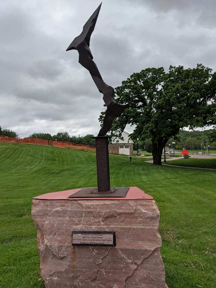 One of the sculptures in Falls Park. It is surrounded by green grass. The metal statue sits on a piece of granite. The sculpture is a line that zig zags towards the sky. The sky is grey with many clouds that are  slightly darker in color than the sky itself.