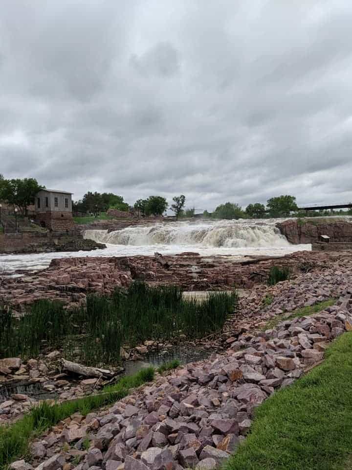 View of the waterfalls in Falls Park. The angle is looking up river at the upper part of the falls. Across the river is the remnants of Queen Bee Mill. On this side of the river are multiple reddish colored rocks and some tall green grasses as the water's edge.
