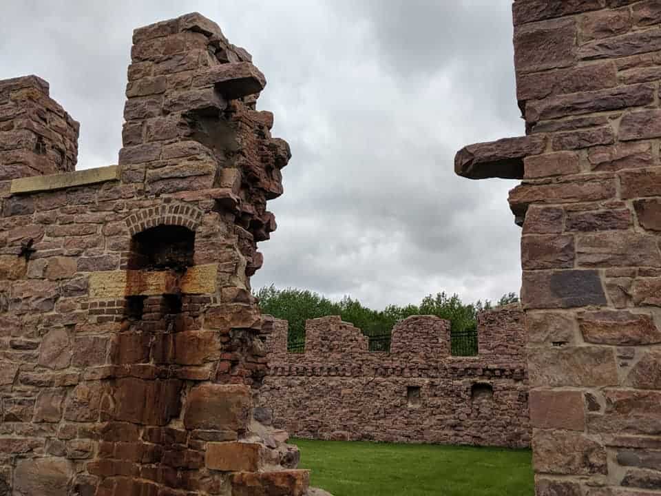 View looking into an opening of the remnants of the Queen Bee Mill in Falls Park in South Dakota. The remnants consist of the exterior brick walls. The middle of area is grass. The walls have battlements spread evenly around the structure.