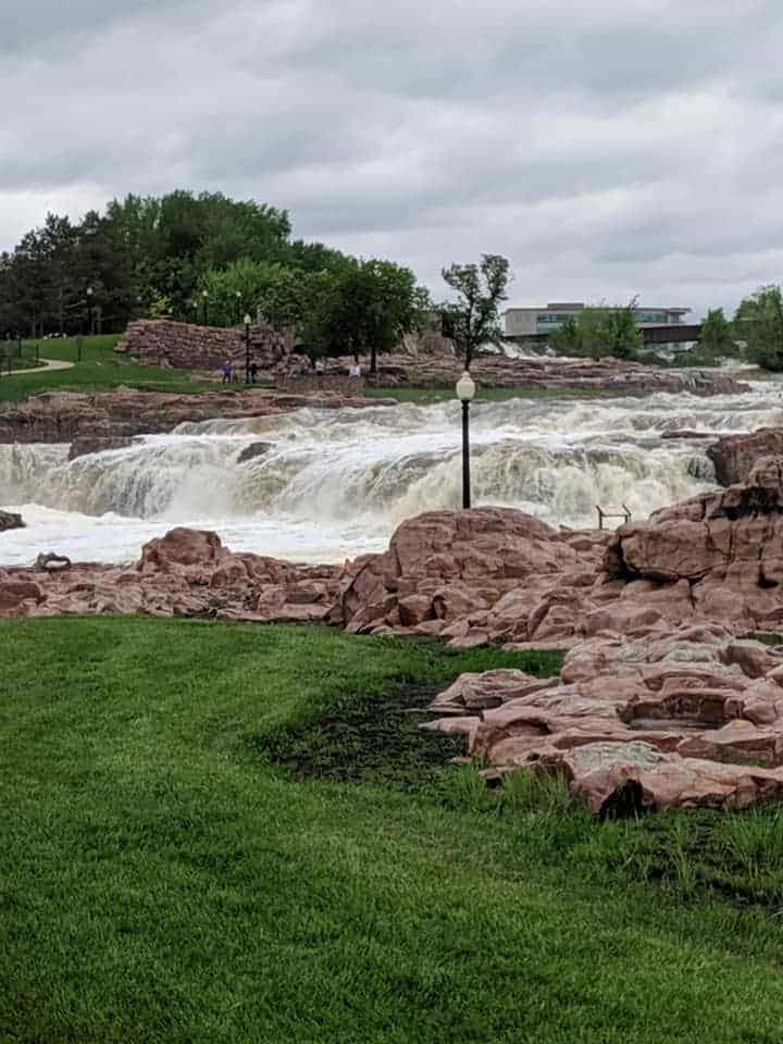 A side view of the waterfalls at Falls Park in South Dakota. There are tons of reddish rocks and boulders along both sides of the river. There is green grass on both sides of the river and rocks. There are decorative lamps throughout the park. You can see a path across the river weaving away and many tall trees in the distance.