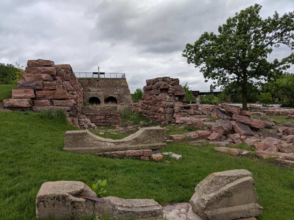 A view of the remnants of the Queen Bee Mill. It is from a side where the wall no longer exists. You can see piles of large bricks on the right side. Further away is what is left of the walls on the left and right side along with the back wall. There is no floor, only grass.