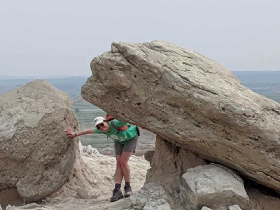 A woman wearing a green t-shirt and grey shorts is bent down and smiling through a passage way between a boulder and another large rock.