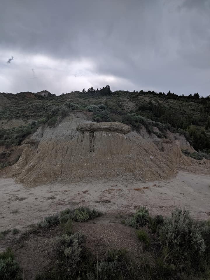 Picture taken looking up from in the Painted Canyon of Theodore Roosevelt National park. The bottom of the hill is bare brown rock. The higher on the hill the more trees and bushes there are. They sky is grey and cloudy due to an incoming storm.