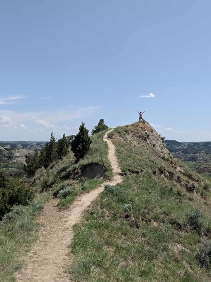 Picture was taken looking up to the top of a hill. There is a dirt path that leads to the top where a woman with a pink shirt has her arms raised to the blue sky. On either side of the path is grass and there are trees on the left side of the path.