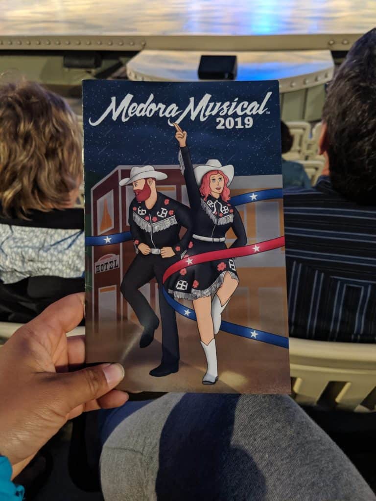 Close up of the brochure for Medora Musical 2019. It has a cartoon man and woman dressed as cowboy and cowgirl with a ribbon that is red and blue with stars on it weaving around them.