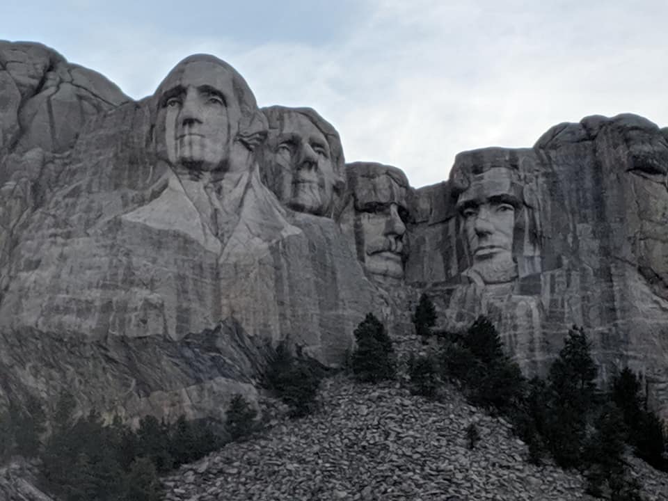 Mount Rushmore with a blue sky in the background. Presidents are seen clearly on the mountain with a line of spruce type trees lined along its base.