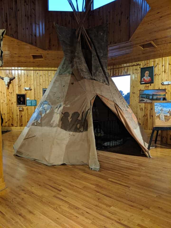 A teepee in the middle of a wooden floor. There are two portraits of Native Americans in the background along with a bull skull and other artwork.