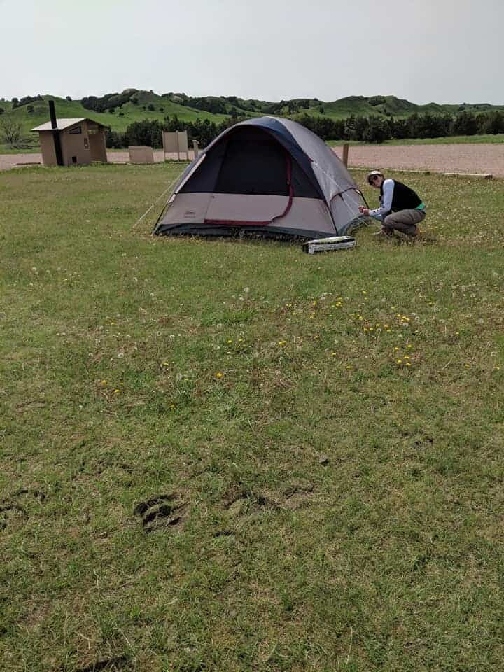 Women crouched down wearing brown pants and a black jacket with white sleeves. She is putting the final touches on a small grey and blue tent at a campsite in Sage Creek Campground. Closer to the photographer are bison hoofprints.