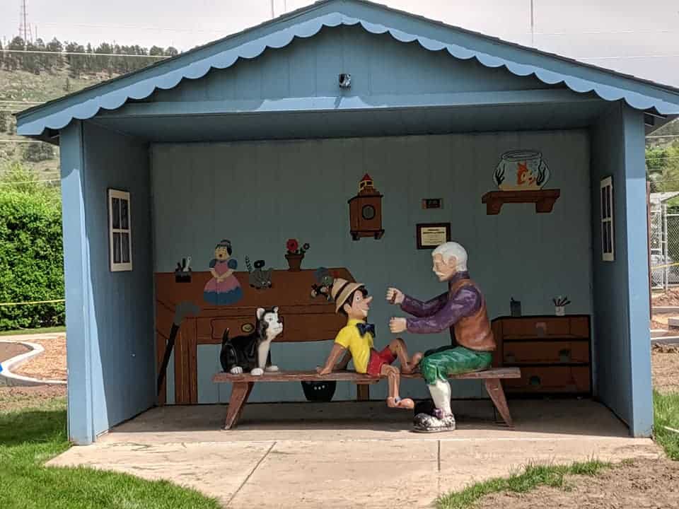 View looking into a room. The wall closest to the photographer doesn't exist. The walls are blue and a desk and dresser are drawn on the back wall. On a long bench in the middle of the room is Pinocchio who is facing Geppetto. Geppetto has his arms reached out to Pinocchio. Behind Pinocchio is Figaro (the black and white cat in the story).  