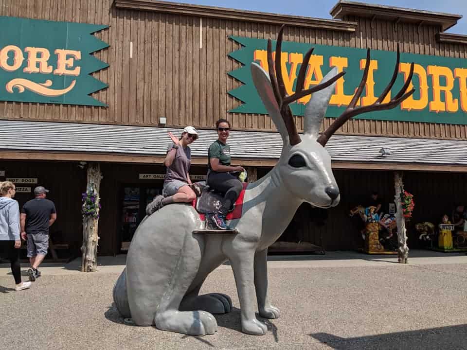 Two women sitting on top of a large grey Jack-o-lope statue with brown antlers. Woman in front has a green t-shirt and grey pants. Woman in back has a grey shirt and khaki shorts.