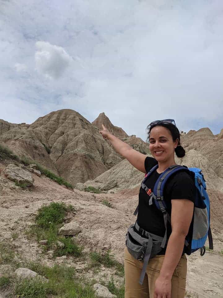Woman wearing a black t-shirt and khaki shorts with a blue back is smiling at camera and point to peak behind her. It is the top of Saddle Pass trail in the Badlands National Park.