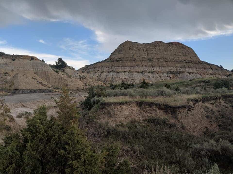 Viewpoint looking up at one of the  large stone hills with lines of color (hues of browns, greys and orange) from the bottom of Painted Canyon . The ground between camera and the hill has grass and small trees. the sky is mostly blue with a grey cloud covering the middle of the sky.