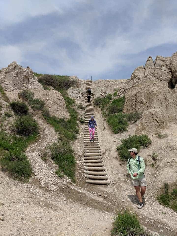 The ladder  on the Notch Trail in Badlands National Park. There are two people almost to the top and a woman with pink pants coming down the ladder about halfway. There is also a man in a green jacket and shorts on the ground looking back at the ladder.