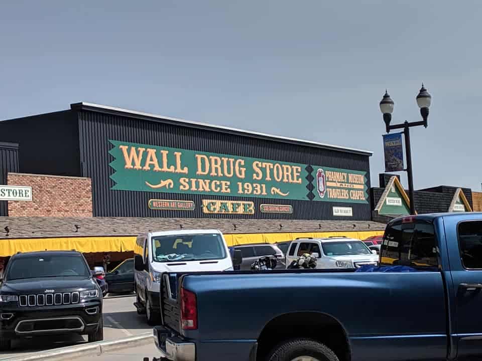 Front of a portion of Wall Drug. There is a large green sign that says "Wall Drug Store Since 1931" in yellow. The top half of building is black, the bottom half is hidden behind many cars in the parking lot.
