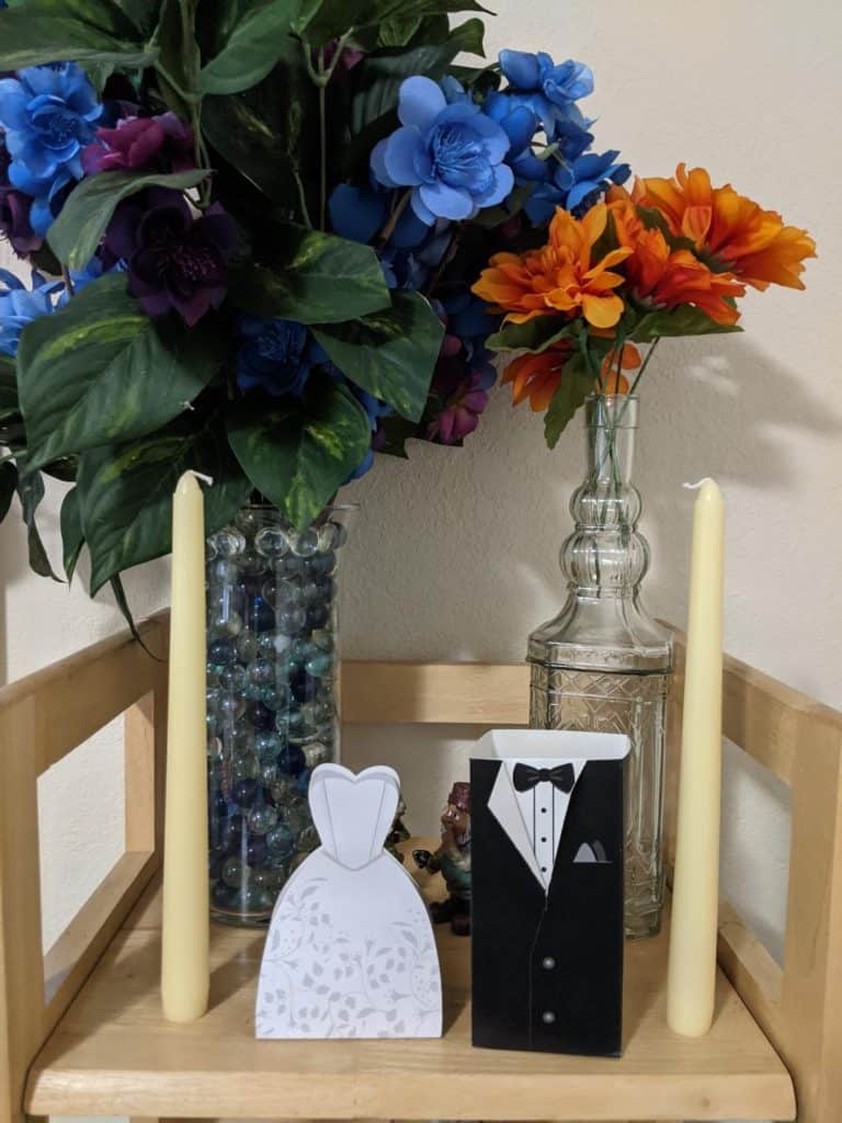 Two bouquets, two candles, and mini cardboard cutouts of a wedding dress and groom tuxedo