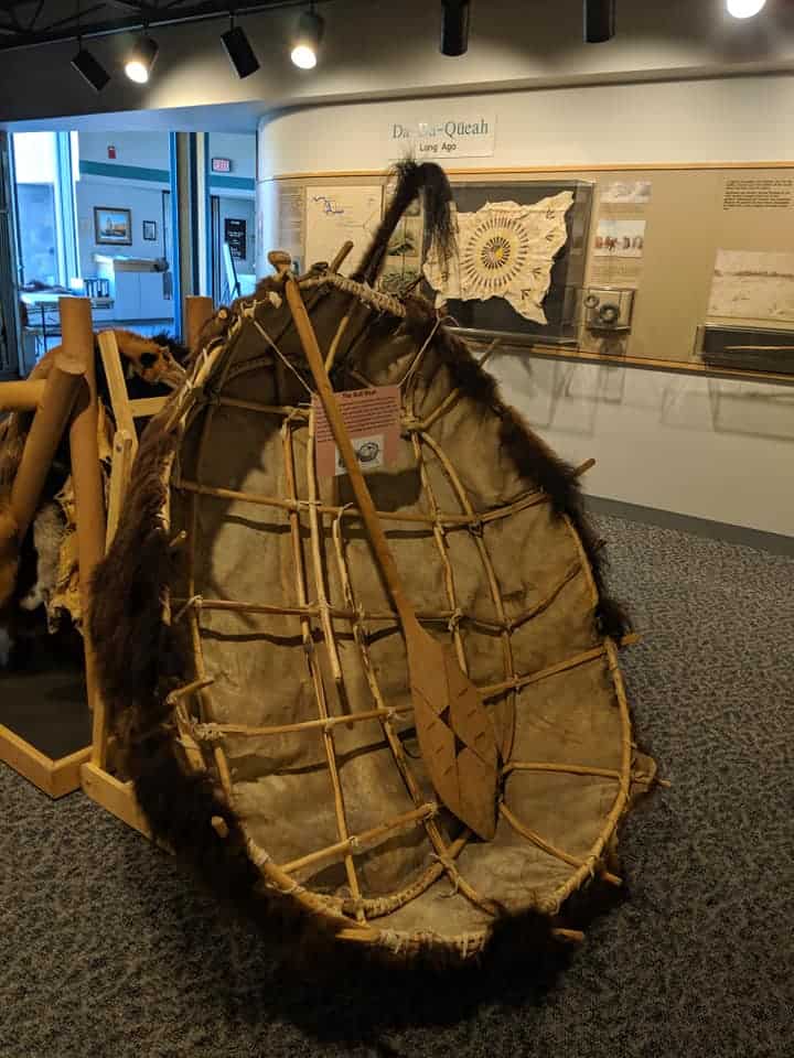 A canoe made from a bison hide, with the hair part on the outside. There is an oar  in the middle of it. Canoe is displayed on a stand in a the Knife River Indian Village NHS visitor Center