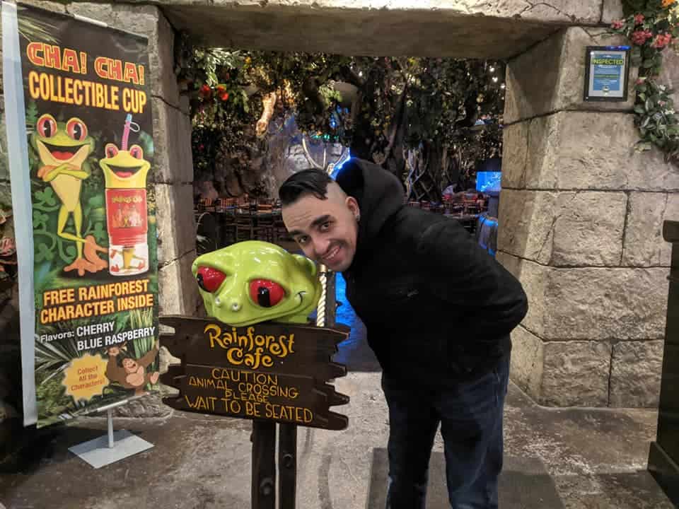 A man in a black jacket leaning towards a green frog with red eyes sitting on top of a Rainforest Cafe sign. They are in the entrance to the restaurant and the man is smiling.