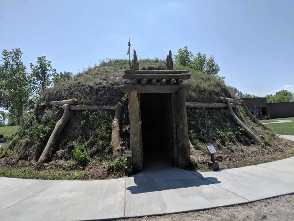 Outside of an Earth Lodge. It is the shape of a mound covered with dirt and grass. The doorway is made of tree trunks.