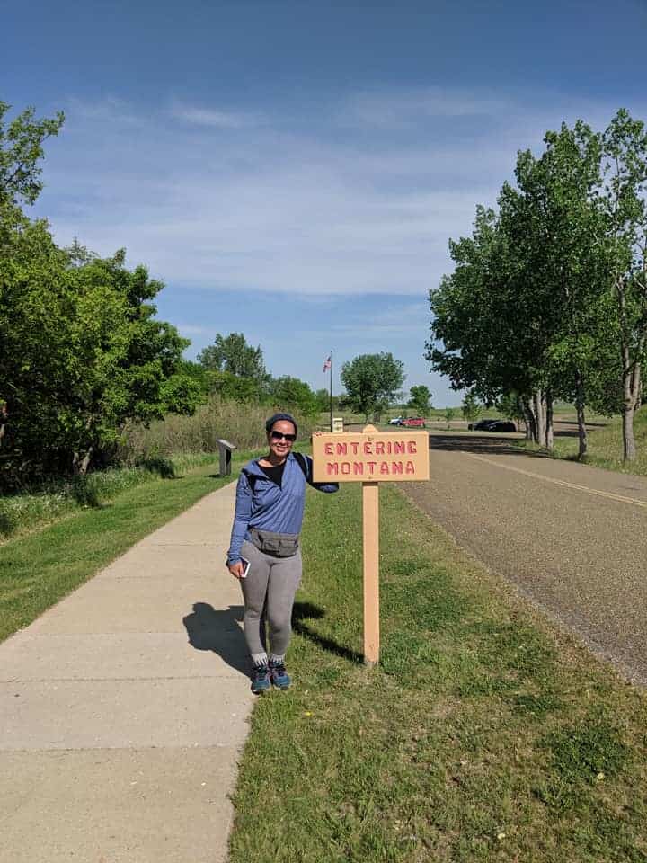 There is woman wearing a blue jacket and grey pants posing next to a brown sign that says "Entering Montana: in red letters. She is standing on a sidewalk that is lined with grass on both sides. The right side is a road and the left side is a row of trees.