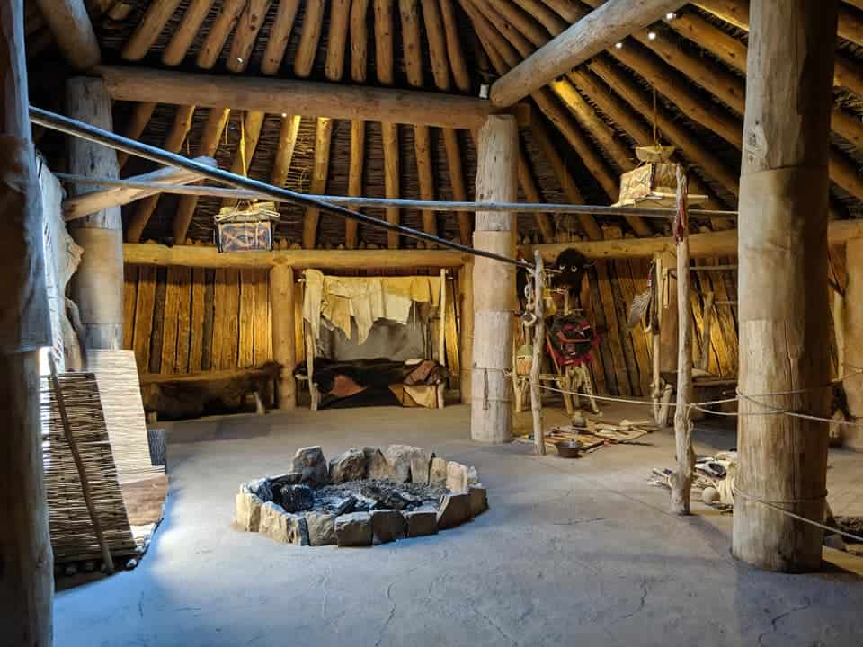 The inside of an Earth Lodge. There is a fire pit in the center and 4 large tree trunks as support beams evenly spaced around the pit. The walls are in a circular shape and made of wooden planks. There is an area with cooking equipment and another where clothes appear to be drying.