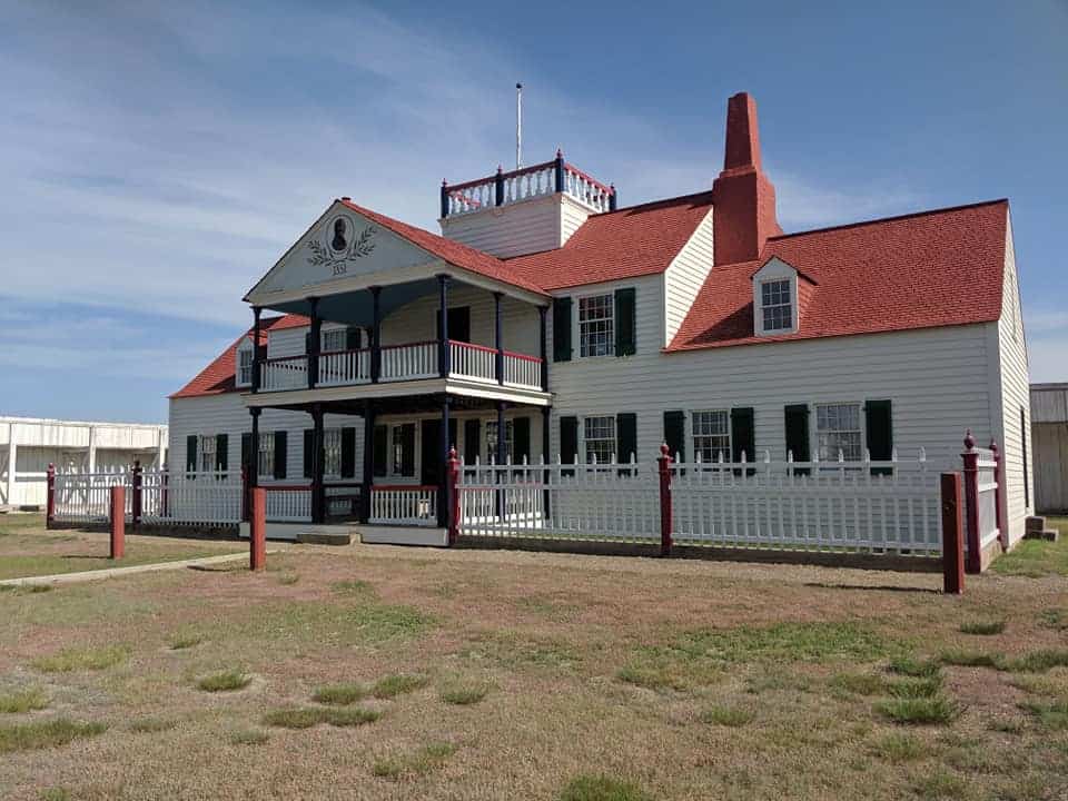 White two story house. With a front porch on both floors. The roof is red and there are black shutters on the 8 bottom windows and two of the tope four. There is a white fend that surrounds the front. It is an old fashioned house; the main house in the Fort Union Trading Post.