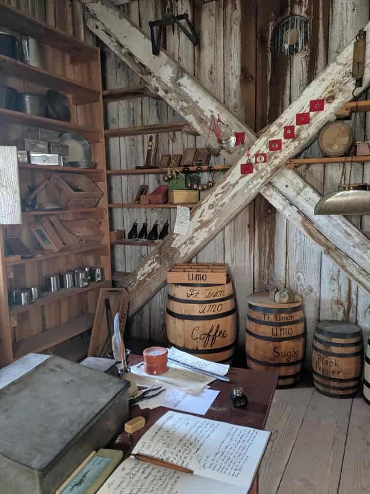 A view of a storage room set up like it would have been when the Fort Union Trading Post was in use. There are 3 barrels that descend in size from left to right on the back wall. The left one says coffee, middle says sugar, and black pepper on the right. There is an open book to record all transactions. The wall on the left have containers with unknown goods in them. The floors and walls are made of wooden planks.