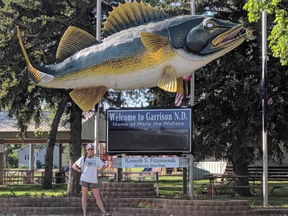 Wally the World's Largest Walley is suspended over a black sign that says "Welcome to Garrison, ND" The Walleye statue is green on top, white belly, and has yellow fins. There is a woman with a white t-shirt and grey shorts smiling at the camera underneath the fish. There are trees and a park shelter behind the fish and woman.