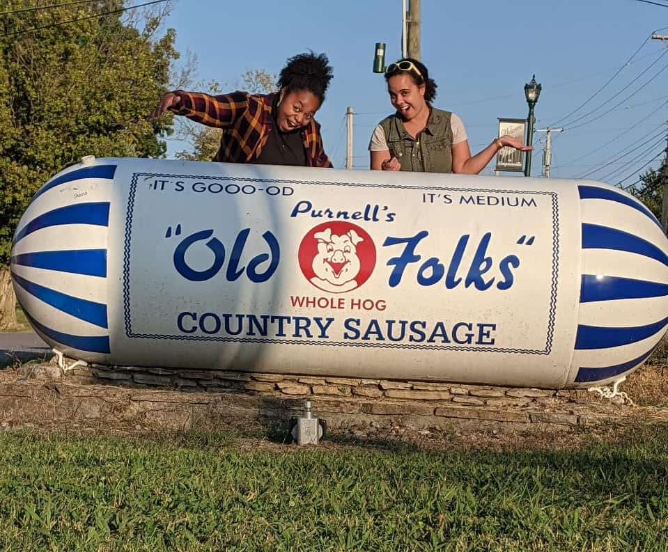 Two women standing behind a fake Country sausage roll that comes up to their mid chest. The sausage roll looks like it is in a white and blue package with "Purnell's Old Folks Whole Hog Country Sausage" written on it.
