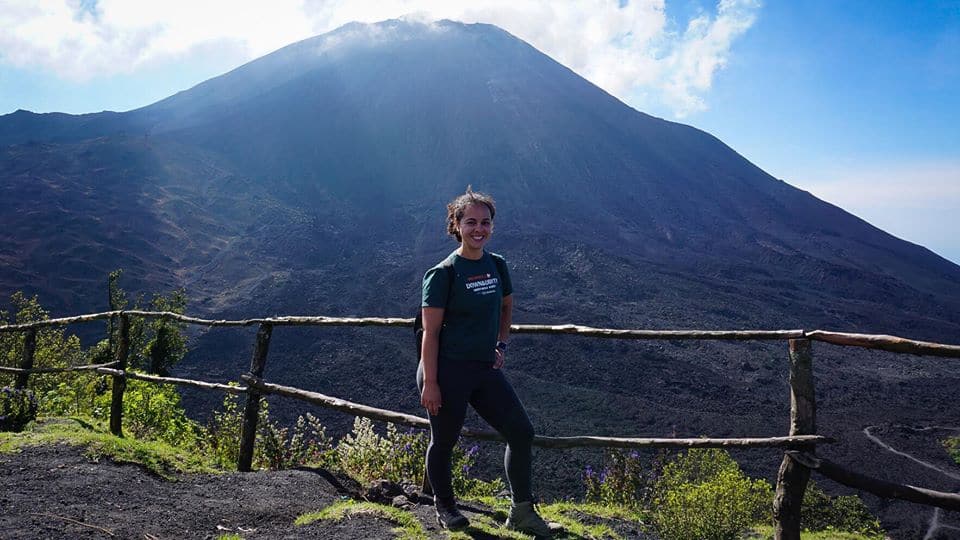 Woman wearing a green t-shirt and black yoga pants standing in front of a simple wooden fence. There is a volcano in the background. Green foliage near her. Sky is blue with clouds and smoke near the tops of volcano.