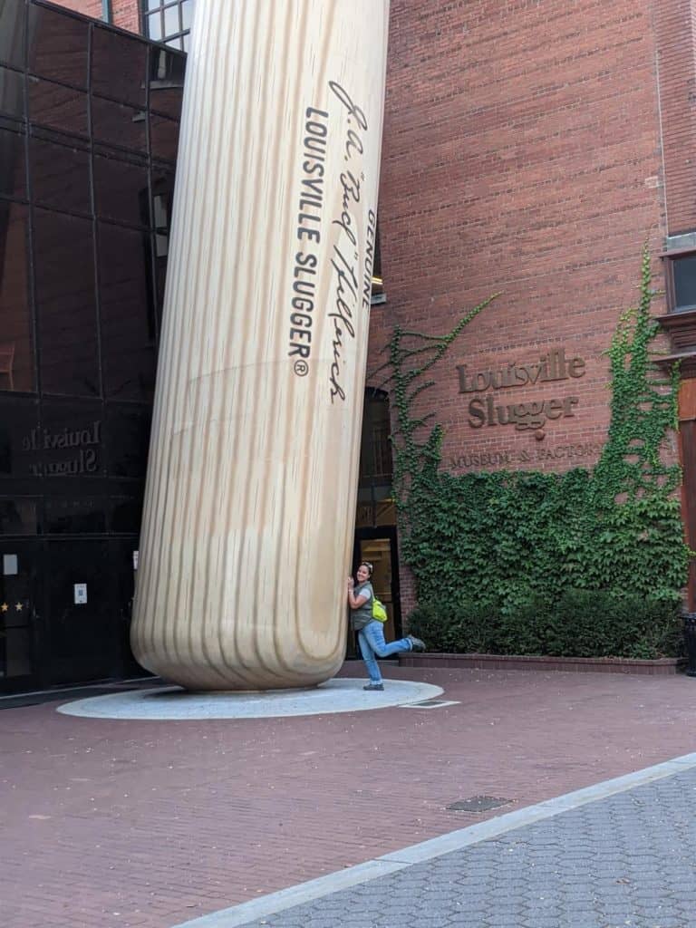 World's Largest baseball in front of the brick Louisville Slugger Museum building. There is a woman wearing jeans and a green vest hugging the bat.