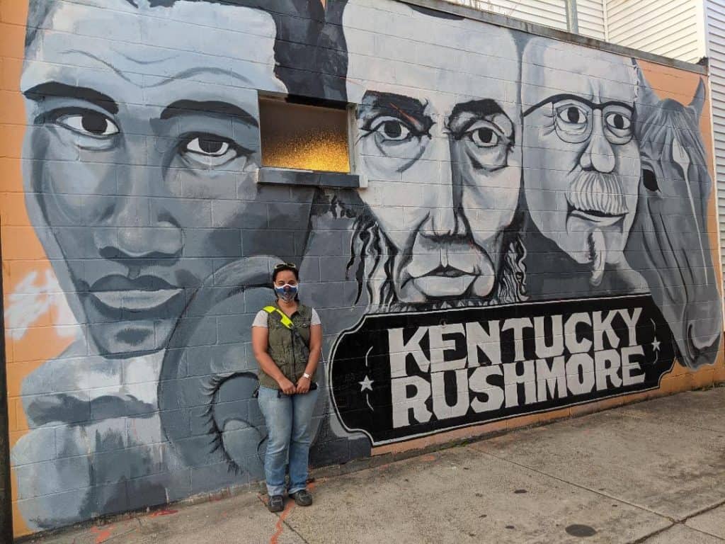 Woman wearing a green vest and blue jeans standing in front of the side of a concrete building with the Kentucky Rushmore mural painted on it. The four figures are painted in black, white, and grey. From Left to Right: Muhammad Ali, Abraham Lincoln, Colonel Sanders and the famous race horse, Secretariat.