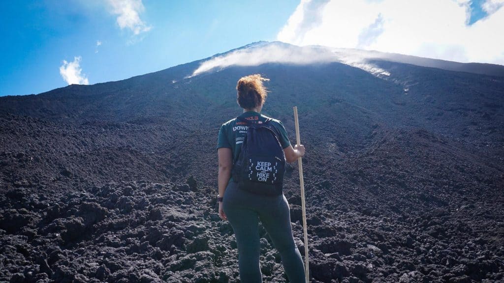 Women wearing a black "Keep Calm and Hike On" backpack is holding a wooden hiking stick facing a volcano with smoke rising from the top. Her back is to the camera.