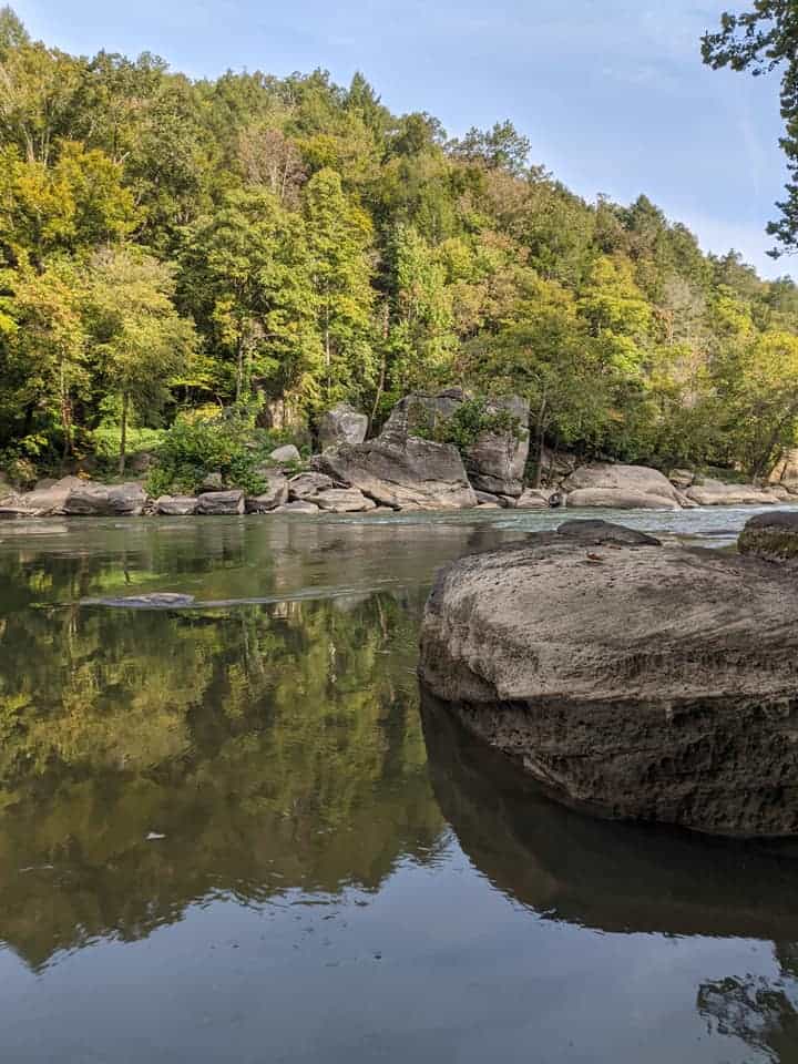View looking across a river in Cumberland State Park with a large boulder to the right. Across the river are more boulders and behind them is a wall of green trees. 