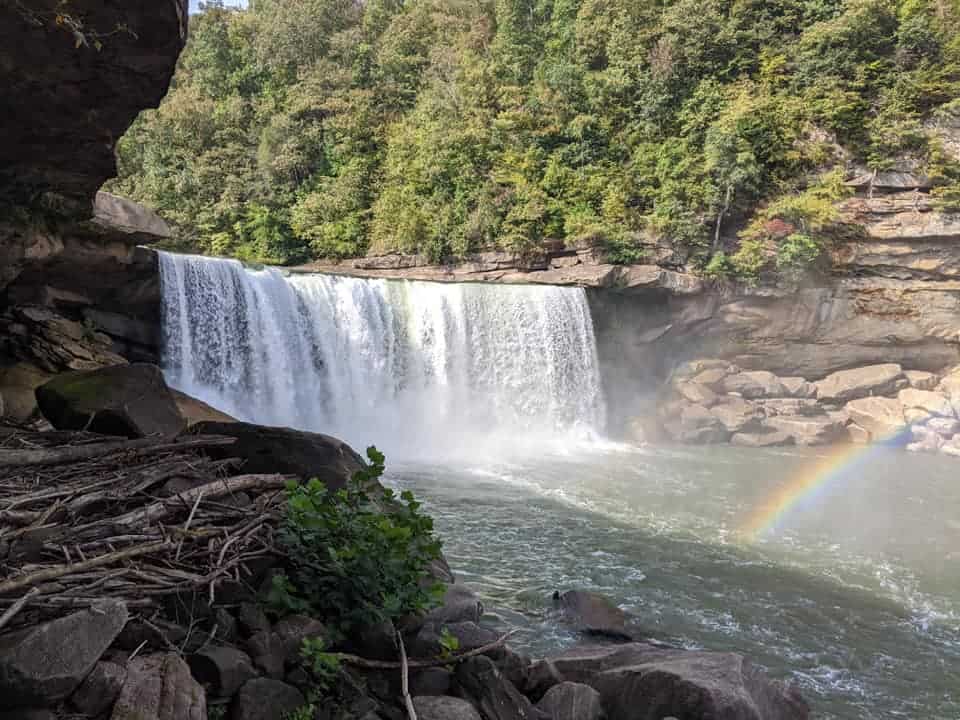 View of Cumberland Falls with a rainbow that appears in the mist. Green forest lies behind the falls.