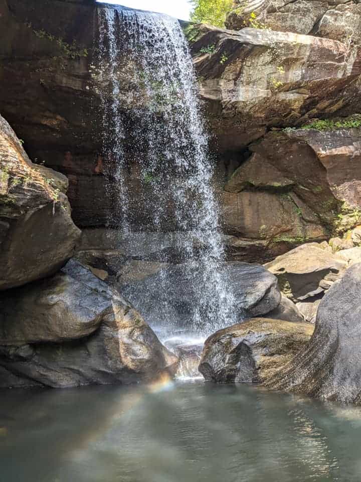 Eagle Falls with the sun shining down on it. A rainbow is reflecting in the pool of water beneath the falls in Cumberland State Park in Kentucky.