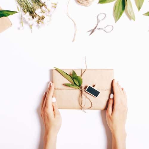 Two hands holding a package wrapped in brown paper. There is a brown cord (ribbon) around it with a green sprig for decoration