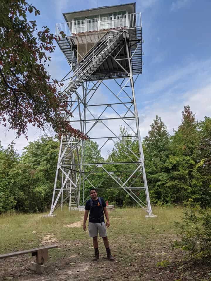 Man standing at the base of Pinnacle Knob Fire Tower in Cumberland State Park. The tower is white in color. There is a forest of green trees in the background.