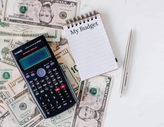 Money (paper bills) are spread out across the left side of a white table. There is a black calculator that is next to a white notepad that says "My Budget" laying on top of the money. The calculator is on the left of the notepad. The notepad is in the middle of the picture. To the right of the notepad is a silver pen.