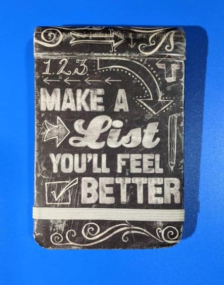 Black notepad with the words "Make a List, You'll Feel Better" written in white. Notepad is laying on a bright blue background.