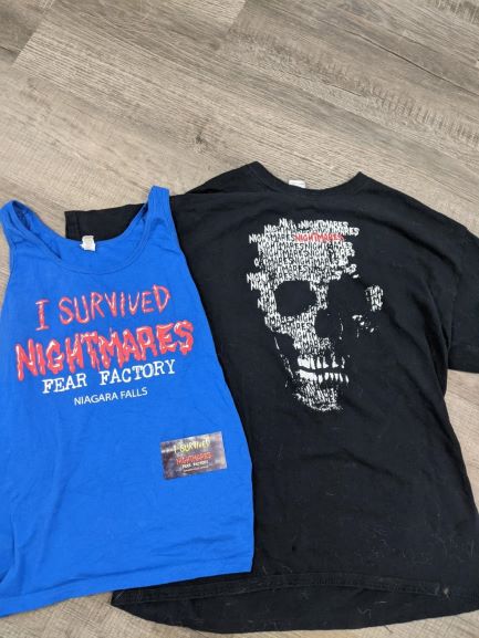 Blue tank top that says "I survived nightmares Fear Factory" in red. It is next to a black T-shirt with similar words in the shape of a skull. There is also a card for the establishment on the blue shirt.