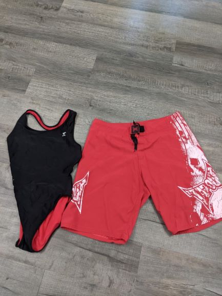 Black 1-piece swimsuit next to a pair of red and white swimming trunks laid out on gray floor.