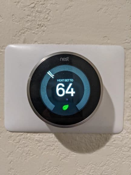 Nest thermostat attached to a cream colored wall. Thermostat heat is set to 64 degrees. There is a tiny leaf beneath the 64 which signifies eco friendly setting.